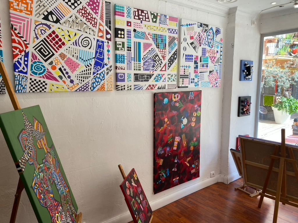 Interior of Galerie Joie De Vivre featuring a collection of vibrant geometric artworks on the wall and additional pieces on easels, with sunlight streaming in from outdoors.