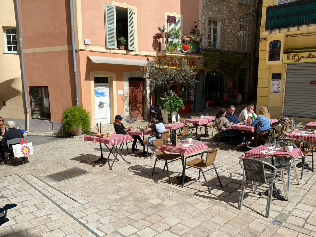 Visitors enjoying the sunny square in front of Galerie Joie De Vivre, with tables set up for dining under the warm Mediterranean sun.